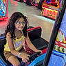 Kimberlee Montero joined the competition — help win amazing prizes! child, girl, curly_hair, glasses, smiling, arcade, arcade_game, racing_game, driving, indoor, amusement_center, entertainment, colorful_lights, neon, seated, yellow_dress, shorts, steering_wheel, portrait, photography