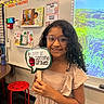 Kimberlee Montero is registered to the contest to win money with this photo: child, girl, school, classroom, second_grade, smiling, portrait, glasses, curly_hair, dress, sign, apple, back_to_school, reading, books, projector, nature, classroom_decor, red_stool, indoor