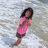 child, smiling, beach, ocean, seawater, waves, surf, foam, shoreline, outdoors, summer, pink_shirt, shorts, curly_hair, wet_hair, standing, portrait, happy, sunlight, vacation