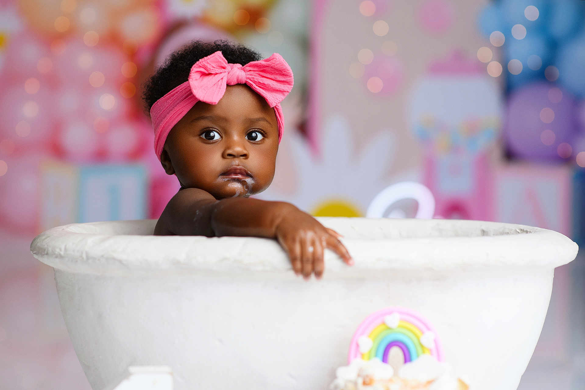 Reign is registered to the contest to win money with this photo: baby, child, pink_bow, headband, tub, milk, cream, face, arm, curly_hair, dark_skin, colorful_background, balloons, blurred_background, cute, innocent, portrait, indoor, decorations, playful