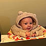 Sutton is registered to the contest to win money with this photo: baby, child, hoodie, teddy_bear, smile, face, seat, high_chair, indoor, curious, cute, clothing, infant, hands, table, wall, person, apparel, expression, young