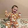 baby, child, onesie, pretzel_pattern, smiling, sitting, carpet, floral_pattern, indoor, happy, cute, person, clothing, footed_pajamas, wall, head, face, expression, young_child, home