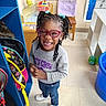 child, girl, glasses, braided_hair, smiling, classroom, toy, backpack, chair, sweatshirt, jeans, sneakers, education, learning, happy, indoor, floor, shelf, colorful, playful