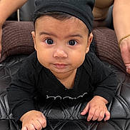 Liam is registered to the contest to win money with this photo: baby, infant, black_hat, bear_ears, black_clothing, curious_expression, indoor, quilted_surface, adult, supporting, hands, face, wide_eyes, cute, portrait, crawling, child, person, skin, headwear
