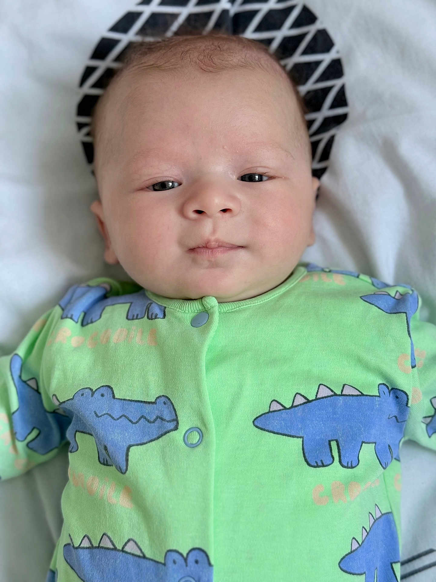 Owen participe au concours pour gagner de l'argent avec cette photo : baby, infant, onesie, green_clothing, crocodile_pattern, blue, face, head, eyes, nose, mouth, skin, lying_down, pillow, white_background, soft_texture, newborn, child, portrait, indoors