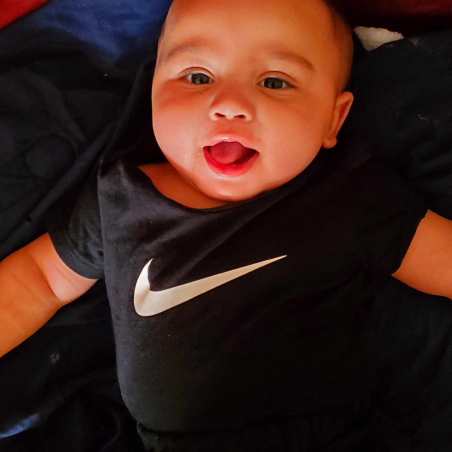 Elijah joined the competition — help win amazing prizes! baby, black_shirt, blanket, blue_blanket, child, clothing, cute, face, happy, head, indoors, infant, laying_down, nike_logo, open_mouth, person, portrait, red_background, skin, smiling