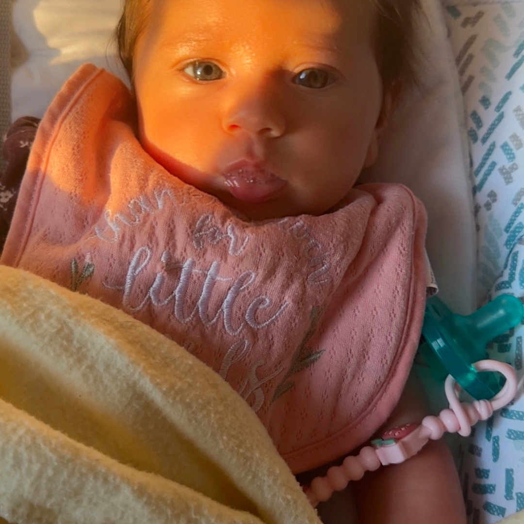 Nathalia is registered to the contest to win money with this photo: baby, blanket, child, close_up, cozy, cute, expression, eyes, face, hand, indoors, infant, newborn, pacifier, peaceful, pink_bib, portrait, skin, sleeping_area, soft_lighting