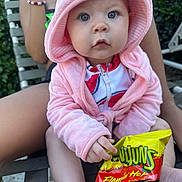 Cecilia is registered to the contest to win money with this photo: baby, child, hooded_jacket, pink_clothing, snack_bag, flamin_hot, outdoor, person, sitting, curious_expression, hand, legs, face, food, snack, bright_colors, greenery, bracelet, relaxation, summer