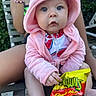 baby, child, hooded_jacket, pink_clothing, snack_bag, flamin_hot, outdoor, person, sitting, curious_expression, hand, legs, face, food, snack, bright_colors, greenery, bracelet, relaxation, summer