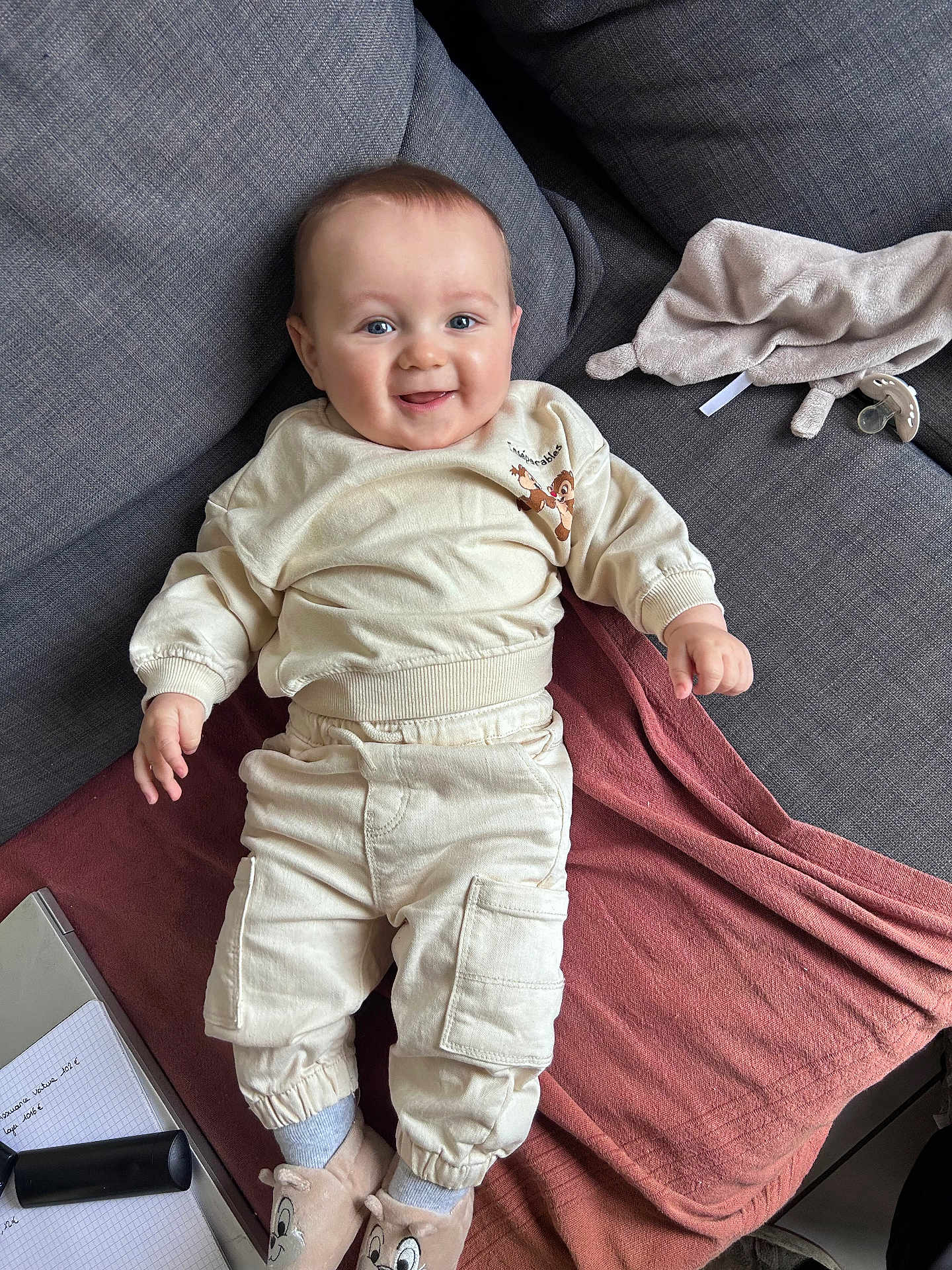 Sacha participe au concours pour gagner de l'argent avec cette photo : baby, infant, smile, happy, couch, sofa, blanket, plush_toy, pacifier, slippers, cream_clothing, pants, sweatshirt, portrait, indoors, relaxed, chubby_cheeks, blue_eyes, hand, furniture