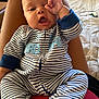 baby, infant, striped_onesie, person, hand_gesture, indoor, bed, legs, lap, cute, face, expression, child, sleepwear, comfort, relaxed, home, soft_lighting, closeup, seated