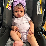 Jerelis is registered to the contest to win money with this photo: baby, infant, car_seat, purple_bow, headband, onesie, serious_expression, child, indoor, person, skin, foot, hand, seat, straps, warning_label, clothing, cute, portrait, young_child