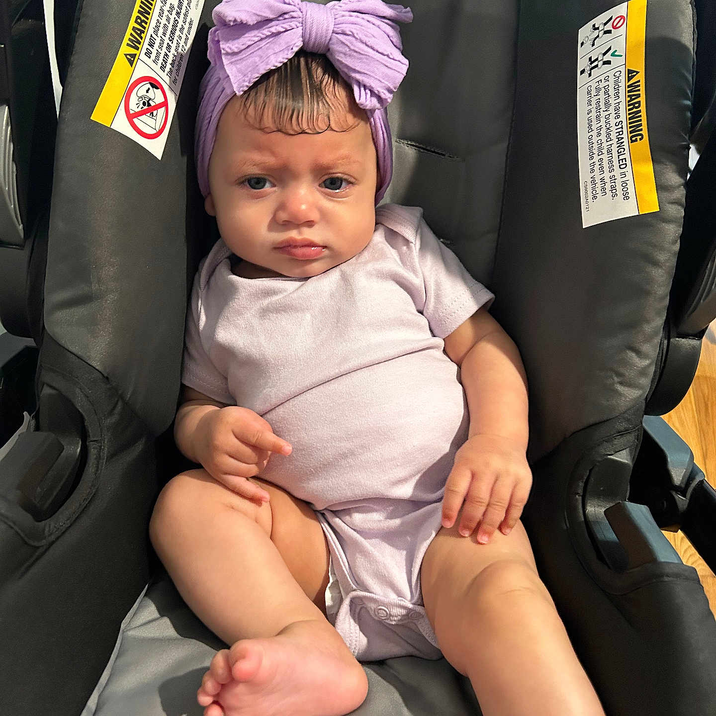 Jerelis is registered to the contest to win money with this photo: baby, infant, car_seat, purple_bow, headband, onesie, serious_expression, child, indoor, person, skin, foot, hand, seat, straps, warning_label, clothing, cute, portrait, young_child