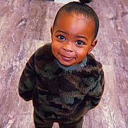 Amiri is registered to the contest to win money with this photo: camouflage, child, clothing, cute, eye_contact, face, floor, hair, happy, indoor, person, portrait, small_child, smiling, socks, standing, toddler, topknot, wood_floor, young_child