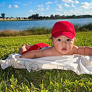 Liam Ortiz is registered to the contest to win money with this photo: baby, child, infant, red_cap, outdoor, grass, blanket, lake, water, sky, clouds, sunny, landscape, lying_down, tummy_time, portrait, face, curious, nature, lakeside