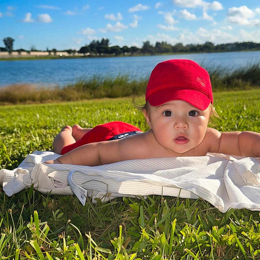 Liam Ortiz is registered to the contest to win money with this photo: baby, child, infant, red_cap, outdoor, grass, blanket, lake, water, sky, clouds, sunny, landscape, lying_down, tummy_time, portrait, face, curious, nature, lakeside