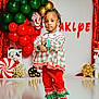 child, girl, holiday_clothing, christmas, festive, balloons, decorations, mickey_mouse, candy_cane, red_pants, plaid, ribbons, braided_hair, greenery, gold_decor, star_lights, snowflake, indoor, portrait, celebration
