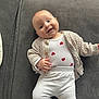 Adalynn is registered to the contest to win money with this photo: baby, infant, smiling, white_pants, heart_pattern, knitted_cardigan, couch, grey_couch, lying_down, happy, child, person, indoors, cute, cozy, casual_clothing, small_hands, head, face, soft_lighting
