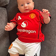 Adalynn is registered to the contest to win money with this photo: baby, child, infant, red_shirt, manchester_united, sportswear, couch, gray_couch, white_shorts, indoors, person, portrait, looking_at_camera, cute, chubby_cheeks, relaxed, comfortable, bright_eyes, shorts, sitting