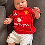 baby, child, infant, red_shirt, manchester_united, sportswear, couch, gray_couch, white_shorts, indoors, person, portrait, looking_at_camera, cute, chubby_cheeks, relaxed, comfortable, bright_eyes, shorts, sitting