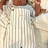 baby, child, infant, portrait, lying_down, bed, bedding, blanket, patterned_background, striped_clothing, overalls, cap, hat, shoes, socks, fashion, adorable, looking_at_camera, indoor, smiling