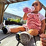 child, toddler, pink_overalls, sitting, outdoor, sunlight, sky, wooden_deck, drinking, bottle, shoes, white_tshirt, pergola, plants, flowers, hand, curious, daylight, casual, young_child