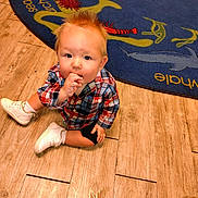 Joe is registered to the contest to win money with this photo: toddler, child, plaid_shirt, white_shoes, wooden_floor, rug, sea_theme, sitting, snack, curious, indoor, cute, spiky_hair, flooring, carpet, baby, person, casual_clothing, footwear, portrait