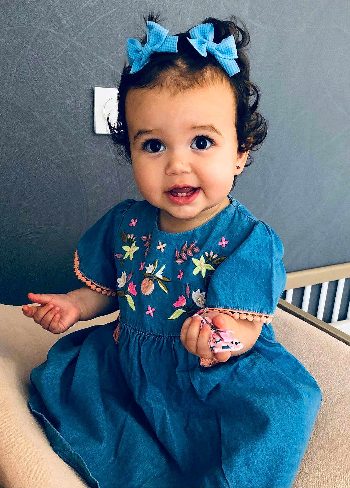 Agata participe au concours pour gagner de l'argent avec cette photo : toddler, child, blue_dress, bows, curly_hair, smile, sitting, indoor, embroidery, hand, face, cute, person, young_child, happy, portrait, clothing, furniture, chair, background