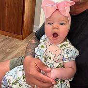 Josephine joined the competition — help win amazing prizes! baby, child, pink_bow, tattoo, adult_hand, indoor, wooden_floor, wooden_furniture, cute, surprised_expression, clothing, patterned_dress, person, holding, portrait, skin, face, sitting, casual, family
