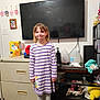 child, girl, smile, purple_striped_dress, boots, television, dresser, clutter, shoes, floor, cabinet, gallon_jugs, bottle, photo_frame, snack_box, shelf, wall, room, portrait, standing