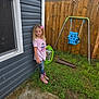 backyard, blue_swing, casual_clothing, child, concrete, daylight, girl, grass, house_wall, jeans, outdoor, pink_boots, pink_bow, pink_shirt, playground, standing, swing, toy, window, wooden_fence
