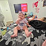 baby, bed, blanket, bow, camouflage, child, clothing, device, furniture, game_console, headband, indoor, person, playing, room, screen, smiling, technology, toddler, wall