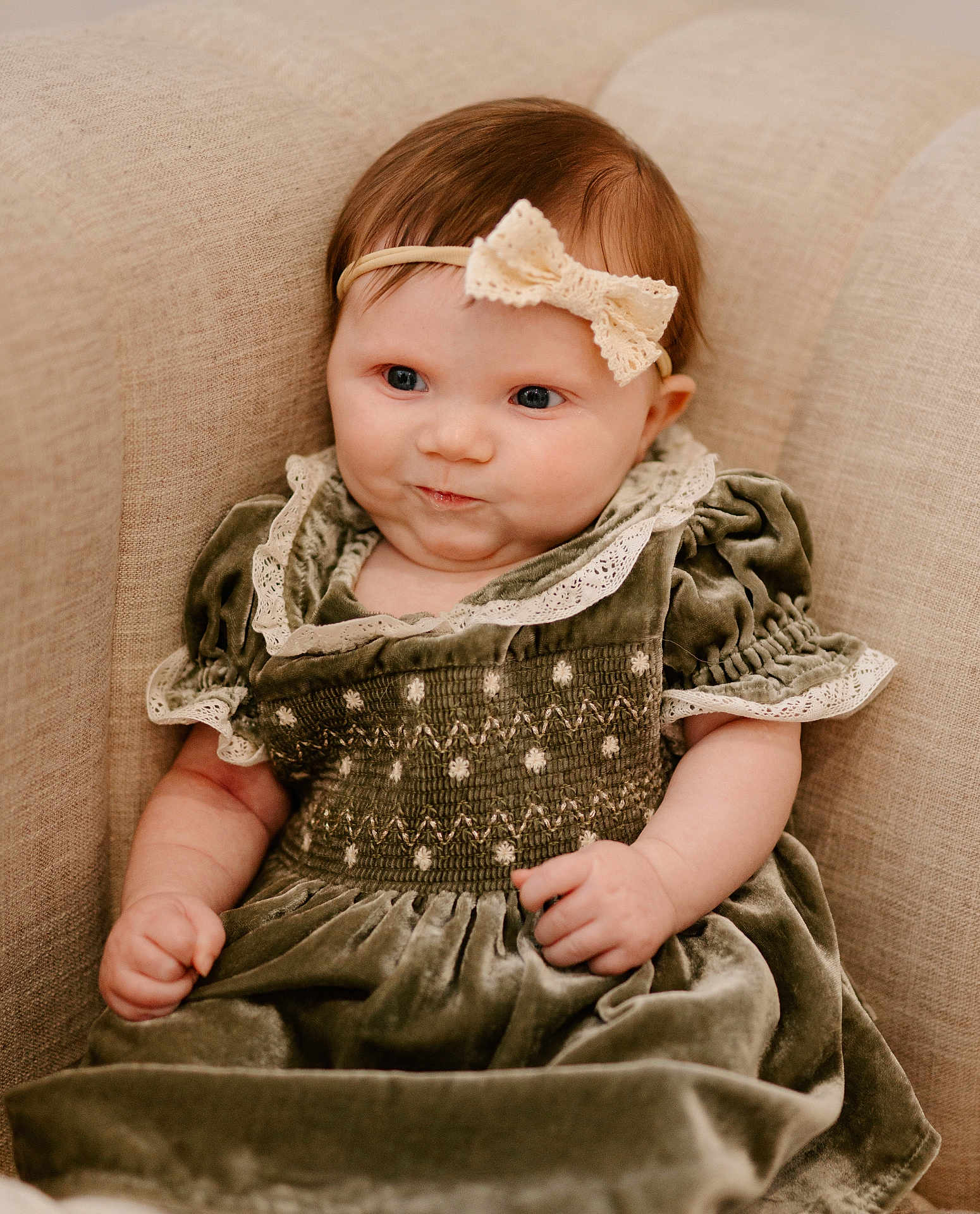 Iyla is registered to the contest to win money with this photo: baby, infant, child, headband, bow, green_dress, velvet, couch, beige, soft_fabric, cute, smirk, indoor, portrait, chubby_cheeks, sitting, neutral_colors, cozy, person, young_child