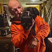 Midnight is registered to the contest to win money with this photo: man, cat, black_cat, orange_hoodie, indoors, kitchen, older_adult, pet, animal, domestic, casual_clothing, facial_hair, holding, shoulder, warm_lighting, cluttered, household_appliance, cupboard, hand, ring