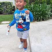 Patrick is registered to the contest to win money with this photo: blue_sweatshirt, boy, bushes, casual_clothing, child, cute, daylight, grass, greenery, happy, mickey_mouse, outdoor, person, shorts, sidewalk, smiling, snack_pack, sneakers, toddler, walking