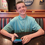 Mikha is registered to the contest to win money with this photo: teenage_boy, smiling, braces, blue_polo_shirt, restaurant, booth, wooden_table, phone, restroom_sign, wall_art, pac_man, indoor, casual, happy, portrait, person, seated, youth, dining, interior