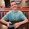 teenage_boy, smiling, braces, blue_polo_shirt, restaurant, booth, wooden_table, phone, restroom_sign, wall_art, pac_man, indoor, casual, happy, portrait, person, seated, youth, dining, interior