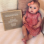 Mackenzie joined the competition — help win amazing prizes! baby, child, infant, pink_bow, dress, cherry_pattern, sign, wooden_sign, text, four_months, pillow, knitted_pillow, white_surface, cute, portrait, sitting, milestone, announcement, indoors, person