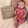 baby, child, infant, pink_bow, dress, cherry_pattern, sign, wooden_sign, text, four_months, pillow, knitted_pillow, white_surface, cute, portrait, sitting, milestone, announcement, indoors, person