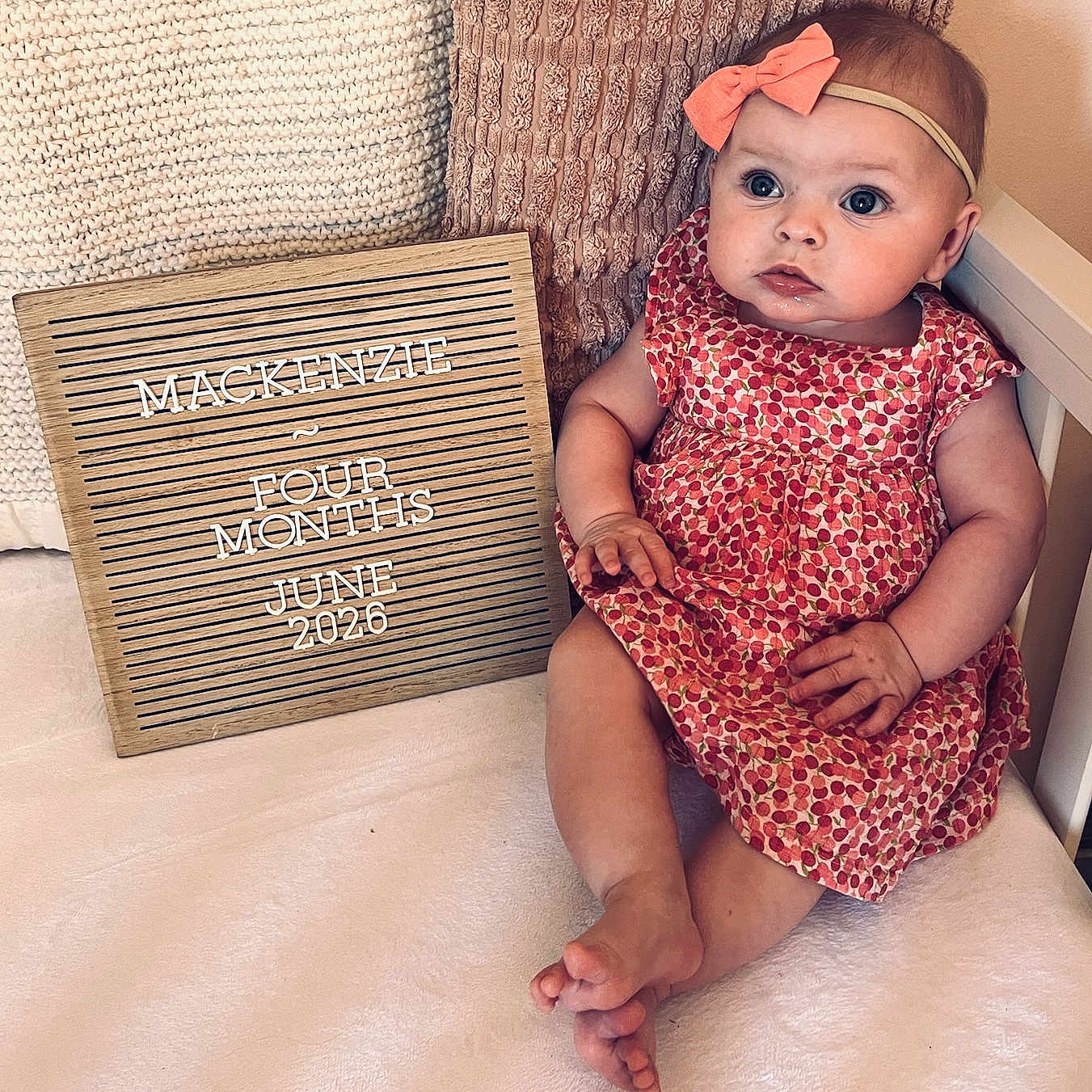Mackenzie joined the competition — help win amazing prizes! baby, child, infant, pink_bow, dress, cherry_pattern, sign, wooden_sign, text, four_months, pillow, knitted_pillow, white_surface, cute, portrait, sitting, milestone, announcement, indoors, person