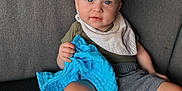 Caiden is registered to the contest to win money with this photo: toddler, child, baby, blue_cloth, gray_couch, shorts, shirt, bib, barefoot, indoor, natural_light, curious, smiling, sitting, person, human, furniture, window, soft_light, casual_clothing