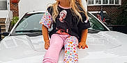 Alex is registered to the contest to win money with this photo: child, blonde_hair, sitting, car, white_vehicle, colorful_clothing, star_pattern, graphic_tshirt, pink_shoes, outdoor, brick_house, red_shutters, american_flag, window, sidewalk, smiling, casual, daytime, young_child, suburban