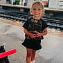 child, girl, smile, train_station, platform, sneakers, black_clothing, shorts, outdoor, railroad_tracks, daylight, casual, standing, young_child, happy, person, urban, background_blur, hair, footwear