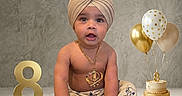 Gurnawab is registered to the contest to win money with this photo: baby, child, turban, birthday, cake, balloons, number_eight, jewelry, bracelet, pants, sitting, celebration, gold, neutral_background, portrait, cute, indoors, happy, milestone, decorations