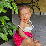 Lylou participe au concours pour gagner de l'argent avec cette photo : baby, child, smiling, sitting, pink_dress, beaded_necklace, barefoot, tiled_floor, green_leaves, plant, outdoor, sunlight, happy, cute, infant, person, portrait, clothing, floor, nature
