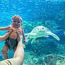 baby, child, person, hands, headband, bow, smile, aquarium, sea_turtle, fish, underwater, water, marine_life, tank, blue, swimming, rocks, coral, nature, cute