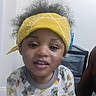 child, toddler, curly_hair, yellow_bandana, pajamas, dinosaur_print, food_print, indoor, face, smiling, person, young_child, cute, casual_clothing, portrait, headwear, expression, home, furniture, drawer