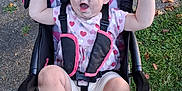 Mara is registered to the contest to win money with this photo: toddler, child, stroller, outdoor, grass, sidewalk, pink_shoes, heart_pattern, raised_arms, happy, messy_face, clothing, seatbelt, black_tray, leaves, daylight, person, cute, playful, nature