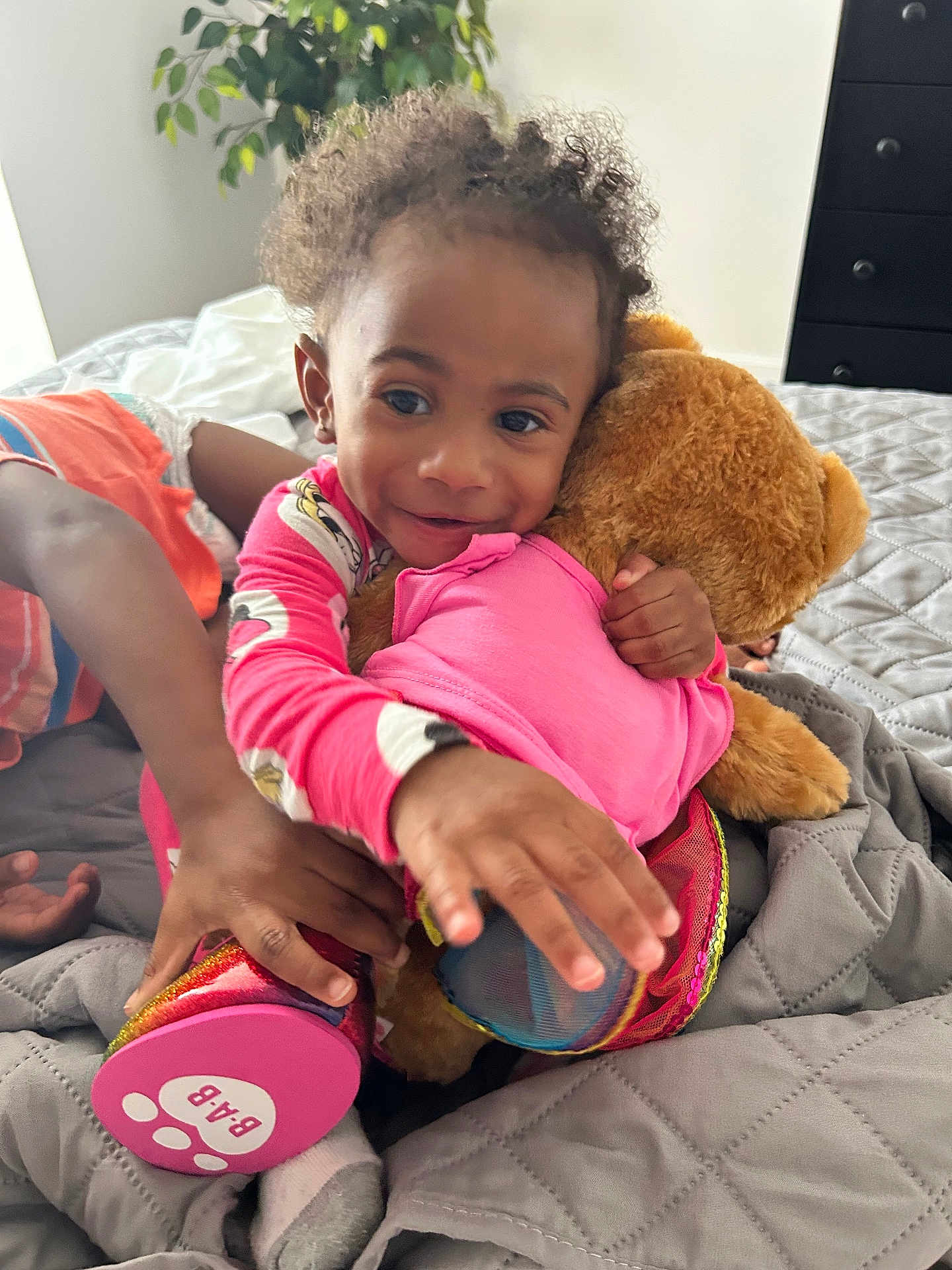 Pheobe joined the competition — help win amazing prizes! toddler, child, teddy_bear, pajamas, bed, blanket, hand, face, smile, toy, plush, furniture, indoor, person, colorful, curly_hair, plant, dresser, footwear, playful