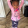 arms_raised, child, curly_hair, girl, happy, indoor, kitchen, patterned_clothing, pink_shirt, playful, portrait, purple_shoes, refrigerator, smiling, sneakers, standing, toddler, trash_can, unicorn, wood_floor